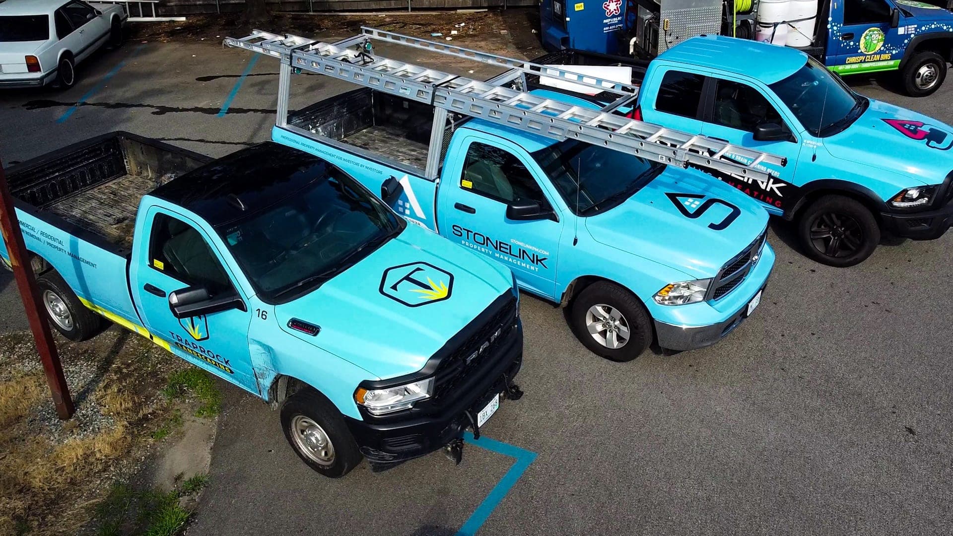 Three light-blue partner trucks parked side by side: a Traprock Landscaping pickup, a Stonelink Property Management truck with ladder racks, and a plumbing and heating pickup.