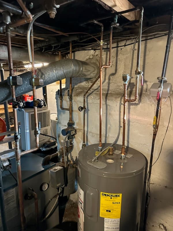 A newly installed water heater with the EnergyGuide label still attached, copper lines overhead, beside the boiler in a stone-foundation basement.